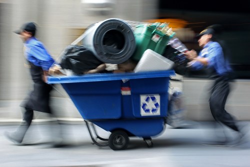A clear, focused photograph of a large blue waste collection bin positioned on a concrete pavement, likely in an urban or industrial area. The bin is filled with various rubbish items including a black plastic bag, a large green waste bin, a rolled-up carpet with visible texture, and a white rectangular object, possibly a plastic container or appliance part. The items are stacked, protruding from the open top of the bin, with some materials leaning outward. The background is blurred, indicating motion, and shows hints of a building and street environment typical of Ilford or surrounding areas. The scene captures the practical aspect of rubbish collection, consistent with services offered by Business Waste Removal Ilford, highlighting their focus on waste disposal and collection in the local community. Bright natural light illuminates the scene, emphasizing the different colors and textures of the waste materials, and the overall composition underscores the importance of efficient rubbish removal for local businesses and residents.