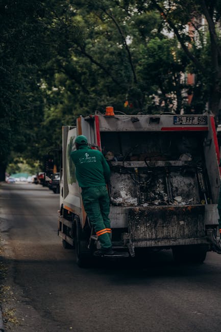 A worker dressed in green safety clothing and a helmet is standing on the back step of a waste collection van parked on a residential street lined with trees. The rear compartment of the van is open, 